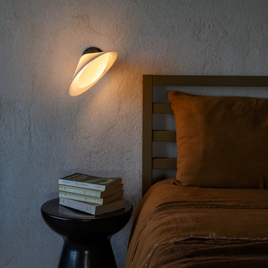DCW Editions Plume wall light, warmly illuminated, angled towards a bed with burnt orange linen bedding