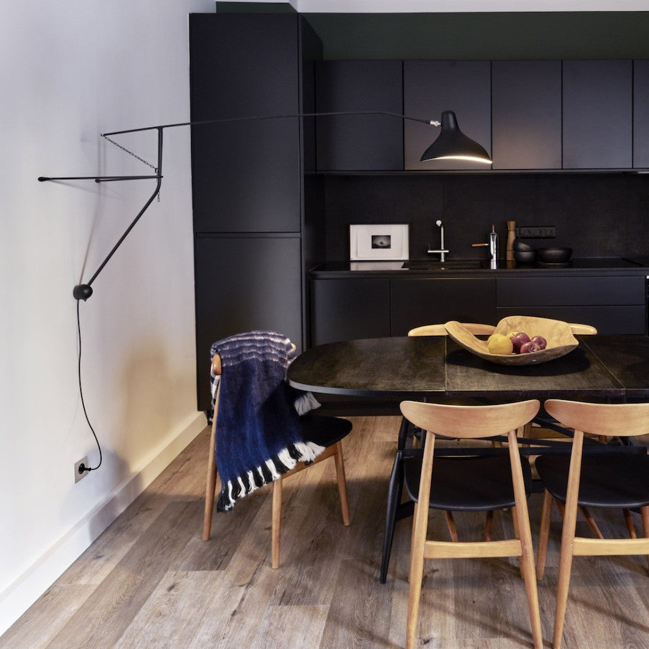 DCW Editions Mantis plug-in wall light BS2 above dark wooden dining table with wooden chairs in front of dark, open kitchen