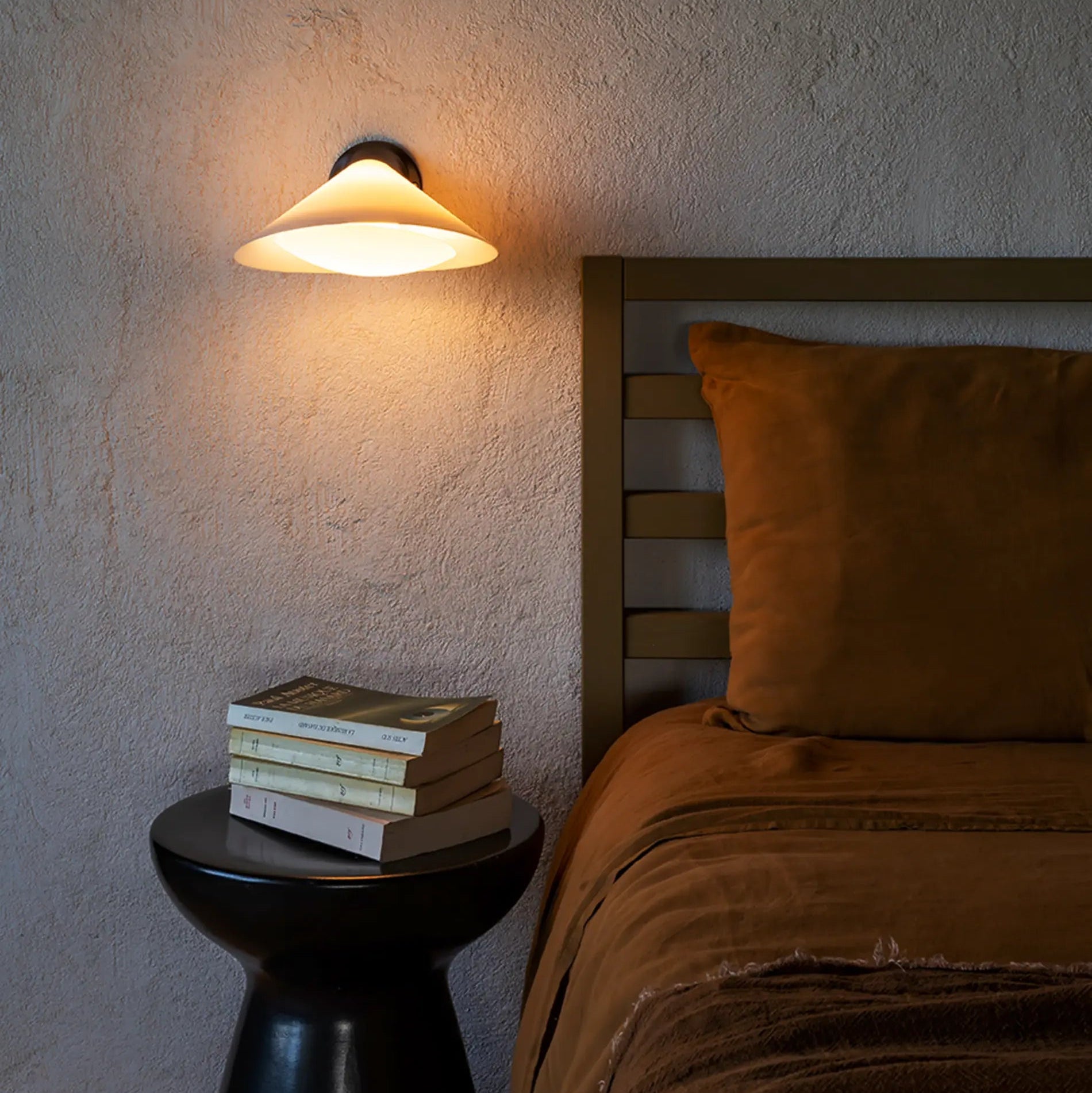 DCW Editions Plume wall light, warmly illuminated, next to a bed with burnt orange linen bedding