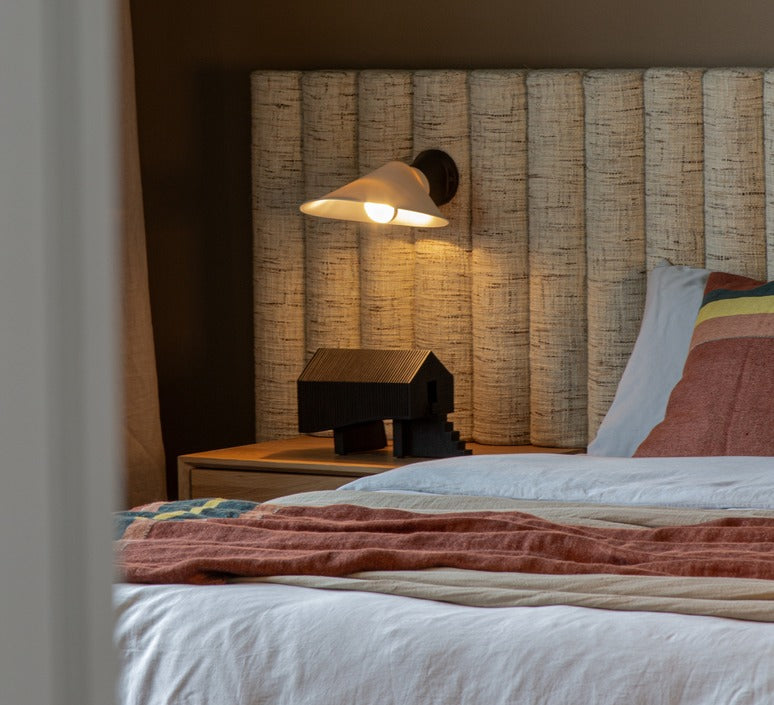 DCW Editions Plume Wall Light over side table next to a bed with a fabric headboard