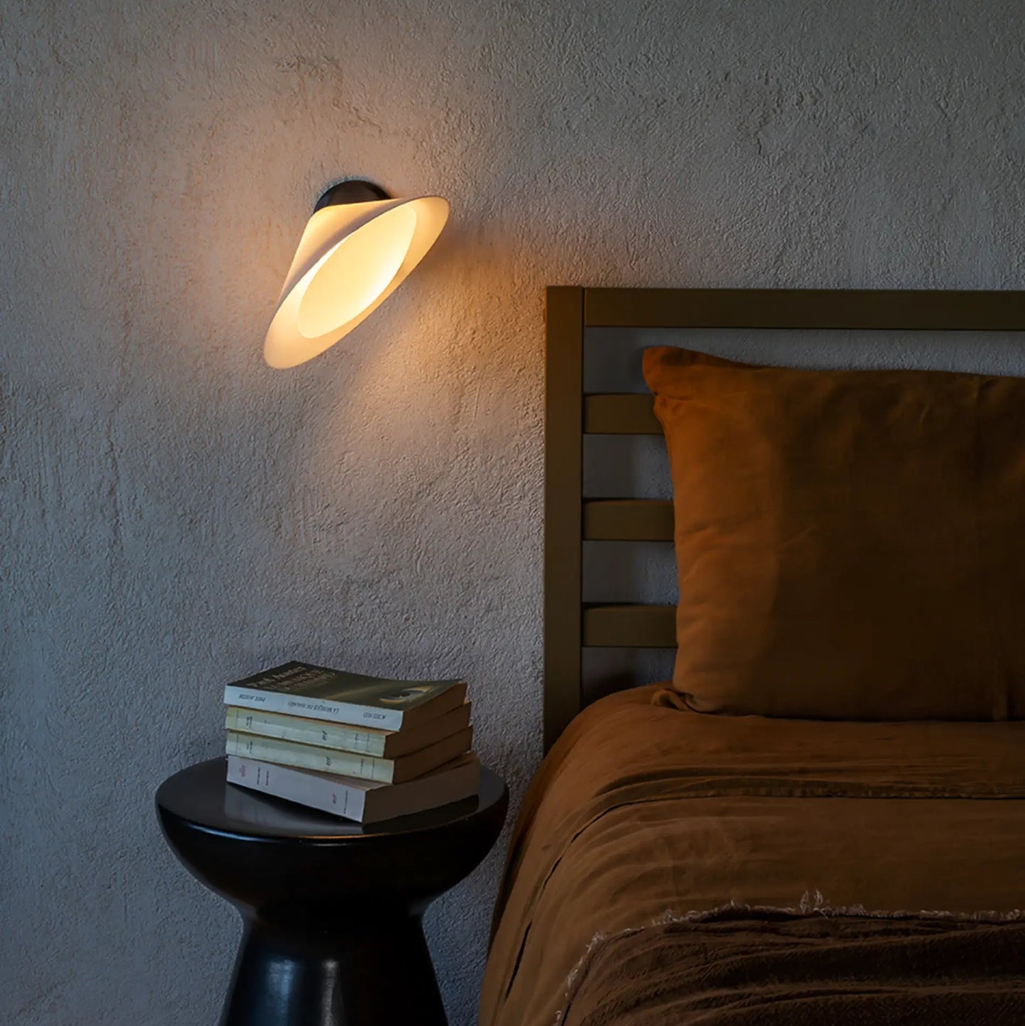 DCW Editions Plume wall light, warmly illuminated, angled towards a bed with burnt orange linen bedding